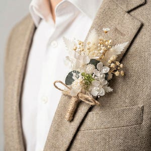 May include: A beige blazer with a white dress shirt and a floral boutonniere. The boutonniere features dried white flowers, greenery, and a twine-wrapped stem. The blazer has a textured appearance.