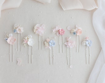 May include: An assortment of floral hair pins in pastel colors, including pink, blue, and white. Each pin features delicate fabric flowers and small embellishments, arranged on silver-toned metal hair pins. The pins are displayed on a neutral background with soft fabric.