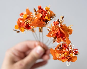 May include: A set of five orange and white dried flower hair pins. The pins are arranged in a bouquet shape and are held in a hand.