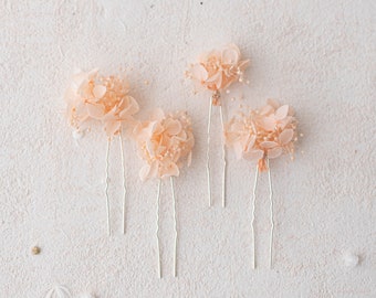May include: Four peach-colored floral hair pins with silver stems. Each pin features a cluster of delicate, dried flowers and small, white accent beads. The pins are arranged on a textured, off-white surface, perfect for bridal or special occasion hairstyles.