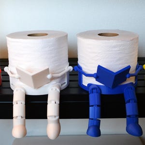 May include: Four toilet paper roll holders, each featuring a figure with articulated legs and arms holding an open book. The holders are colored red, white, blue, and yellow, and each supports a standard white roll of toilet paper.