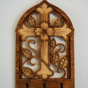 May include: A wall-mounted wooden key holder featuring an arched frame with a central cross design. The cross is adorned with a carved flower and leaf motifs. The piece has a two-tone brown finish and includes three black metal hooks at the bottom.