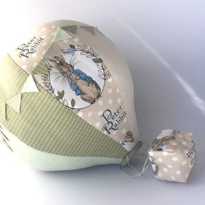 Pode incluir: Um brinquedo de balão de ar quente de tecido com um design de Peter Rabbit. O balão é bege e verde com uma ilustração de Peter Rabbit e as palavras "Peter Rabbit". Um pequeno brinquedo em forma de cubo está anexado.