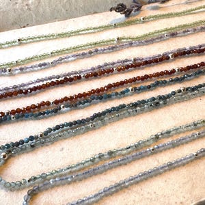 May include: An assortment of beaded necklaces in various colors, including green, purple, brown, and blue. Each necklace features small, faceted beads and silver-colored accents. The necklaces are displayed on a light-colored surface.