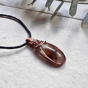 May include: A handcrafted pendant necklace featuring a clear, oval-shaped stone with brown inclusions, encased in copper wire. The pendant hangs from a black cord. The jewelry is displayed on a textured, off-white surface.