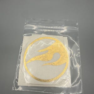May include: A clear plastic bag containing several gold vinyl stickers. Each sticker features a circular design with a flame-like pattern inside. The stickers are on a white backing.