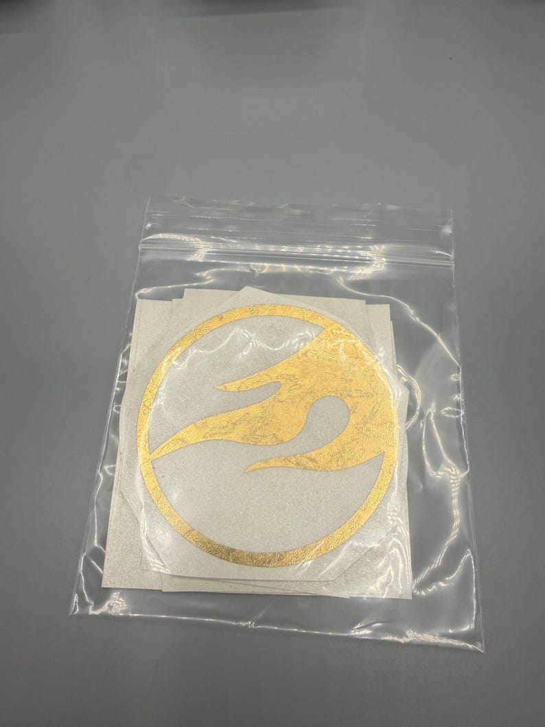 May include: A clear plastic bag containing several gold vinyl stickers. Each sticker features a circular design with a flame-like pattern inside. The stickers are on a white backing.