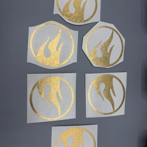 May include: Several gold flame-shaped stickers in different sizes. Each sticker has a flame design within a circular or geometric border. The stickers are on a white backing, with a gold metallic finish, and are set against a grey background.
