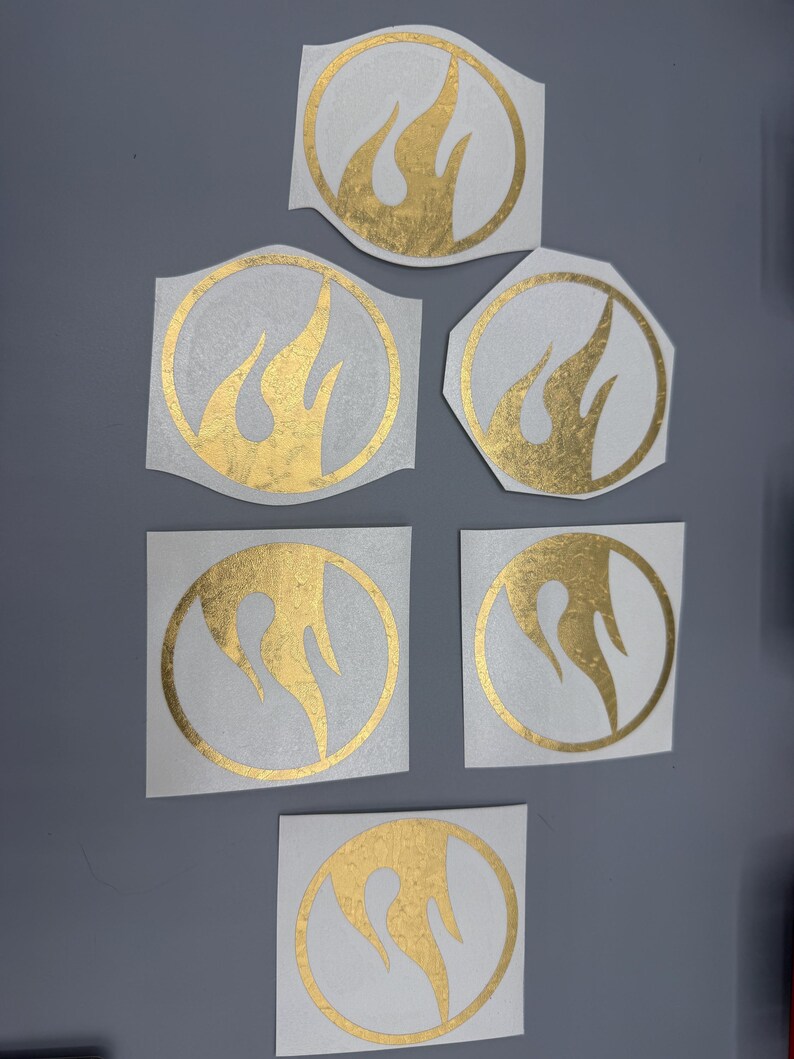 May include: Several gold flame-shaped stickers in different sizes. Each sticker has a flame design within a circular or geometric border. The stickers are on a white backing, with a gold metallic finish, and are set against a grey background.