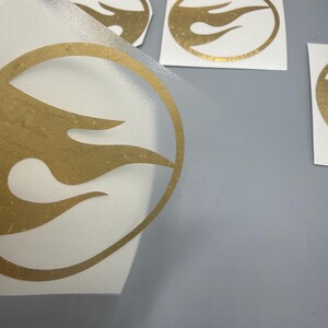 May include: Gold-coloured flame logo stickers on a white background. The stickers are in various sizes and shapes, with a clear backing. The logo features a stylized flame design within a circular outline.