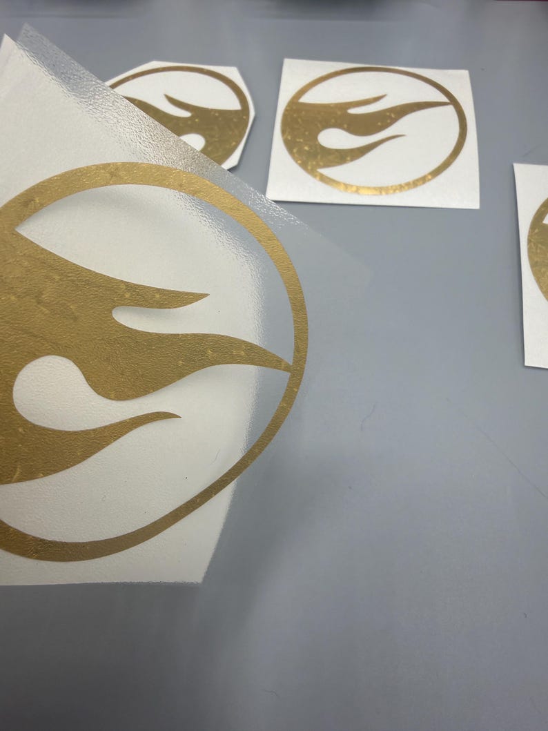 May include: Gold-coloured flame logo stickers on a white background. The stickers are in various sizes and shapes, with a clear backing. The logo features a stylized flame design within a circular outline.