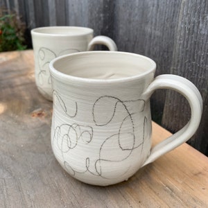May include: Two white ceramic mugs with black scribbled line designs. The mugs have large handles and a textured surface. One mug is in focus, the other is slightly blurred in the background. The mugs are on a wooden surface.