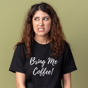 May include: A woman with curly brown hair is wearing a black t-shirt with the text "Bring Me Coffee!" in white lettering.