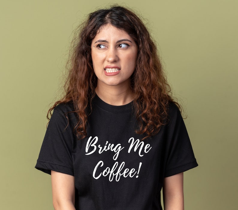 May include: A woman with curly brown hair is wearing a black t-shirt with the text "Bring Me Coffee!" in white lettering.