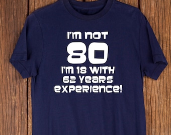 Puede incluir: Camiseta azul marino con el texto blanco: "I'M NOT 80 I'M 18 WITH 62 YEARS EXPERIENCE!". La camiseta está colgada en una percha color cobre sobre un fondo de madera. El texto es en negrita, sin serifa.