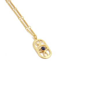 Puede incluir: Un collar de cadena dorada con un colgante que presenta un ojo estilizado con una piedra preciosa azul oscuro en el centro. El colgante está rodeado por un diseño delicado e intrincado.