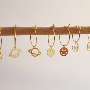 Peut inclure: Un ensemble de boucles d'oreilles créoles dorées avec différents pendentifs, dont un trèfle à quatre feuilles, une planète, un smiley, un cœur et un chat.