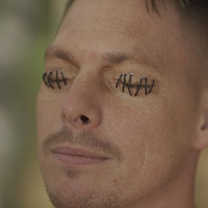 May include: Close-up of a person's face with closed eyes. The eyelids are stitched shut with black thread, creating a stark contrast against the skin. The person has a short haircut and a mustache.