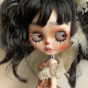 Può includere: Una bambola con i capelli neri raccolti in due codini, indossa un colletto di pizzo bianco e un vestito marrone con bordi di pizzo bianco. La bambola ha il trucco dipinto, comprese le guance rosse e grandi occhi bianchi con eyeliner nero.