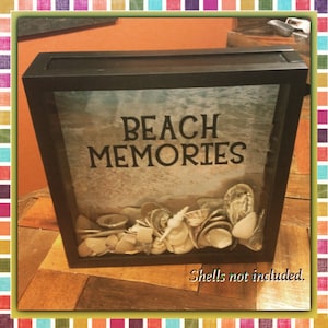 May include: Black shadow box with the words "BEACH MEMORIES" and a beach scene background. The box is filled with various seashells. The text "Shells not included" is at the bottom.