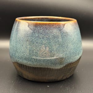 Op de afbeelding: Een kleine, ronde keramische pot met een donkerbruine rand en een blauwe glazuur. De glazuur heeft een gespikkelde uitstraling en een donkerdere bruine band rond de bodem.