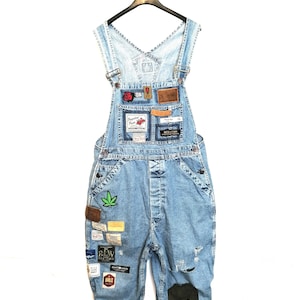 May include: Light blue denim overalls with multiple patches and a green marijuana leaf patch. The overalls have a single button closure and are distressed with rips and tears.