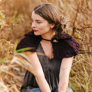 May include: A woman wearing a dark purple knitted shawl with a brown leather strap and a gray dress. She is sitting in a field of tall grass.