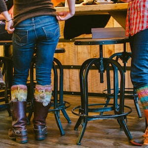Puede incluir: Tres personas con polainas de punto de colores y botas se encuentran frente a un mostrador de madera con taburetes de bar de metal. Las polainas son una mezcla de rosa, azul, verde y amarillo.