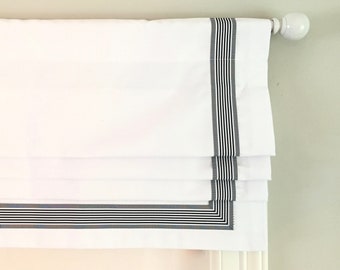 May include: White roman shade with black and gray striped trim. The shade is raised, showing the folds of the fabric.