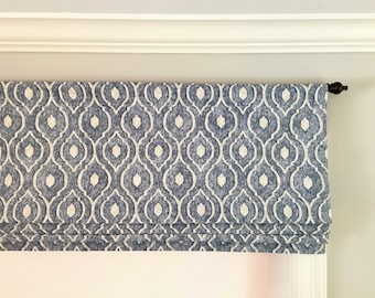 May include: Blue and white patterned Roman shade. The fabric features a repeating quatrefoil design in white against a blue background. The shade is mounted on a dark brown rod and is partially raised, revealing the window frame.