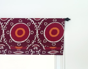 May include: A burgundy roman shade with a white and orange geometric pattern. The shade is mounted on a white window frame.