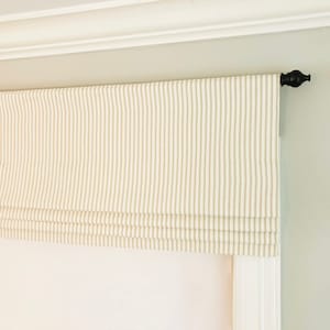 May include: A white and beige striped roman shade with a black finial on the right side.
