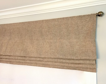 May include: A neutral-toned Roman shade window treatment. The shade is made of a textured fabric in a light brown color. The shade is mounted above a window and has a decorative finial on the rod.