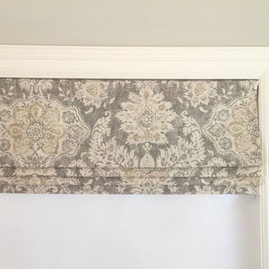 May include: A gray and white patterned roman shade with a white trim. The shade is raised, showing the fabric folds.