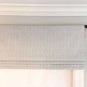 May include: A white roman shade with a small white pattern on a gray background. The shade is mounted to a white window frame.