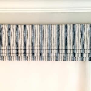 May include: A white and blue striped roman shade with a valance, mounted on a white window frame.