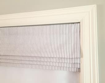 May include: White roman shade with thin black stripes. The shade is mounted inside a white window frame.