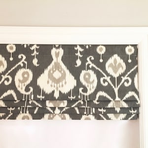 May include: A white window frame with a gray and white patterned roman shade. The shade has a geometric design with white and gray swirls.