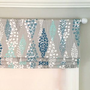 May include: A gray roman shade with a blue and white abstract pattern. The shade is made of a textured fabric and has a white trim.