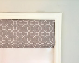 May include: A gray Roman shade with a white geometric pattern, installed within a white window frame. The shade is partially lowered, revealing its folded design. The wall is painted a light gray color.