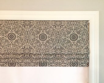 May include: A black and white patterned roman shade with a white trim. The shade is pulled up to reveal a white window.
