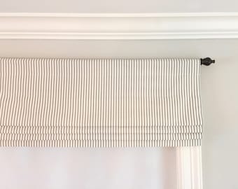 May include: A white roman shade with thin gray stripes hangs from a brown metal rod. The shade is raised, revealing a white curtain underneath.