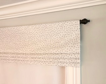May include: A white roman shade with a gray and white speckled pattern. The shade is mounted on a black metal rod.