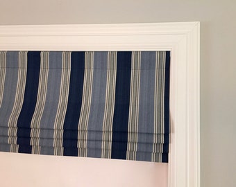 May include: A blue and white striped Roman shade is mounted inside a white window frame. The fabric features vertical stripes in navy, light blue, and gray. The shade is partially raised, showing the folded bottom.