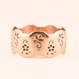 May include: A rose gold ring with a scalloped edge and intricate cut-out designs. The ring features a repeating pattern of grape clusters and floral motifs, creating a delicate and ornate appearance. The ring is a wide band.