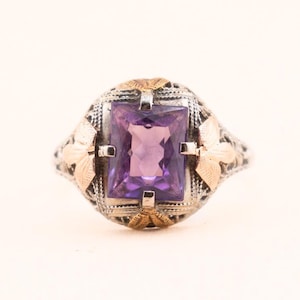May include: A ring featuring a rectangular, faceted purple amethyst gemstone set in a silver-toned metal band with intricate filigree detailing and small, gold-toned accents on the sides of the central stone setting.
