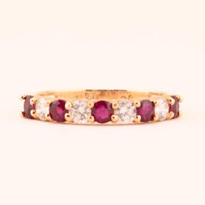 Peut inclure: Une bague en or ornée de pierres précieuses rouges et claires alternées. La bague est sertie d'une série de rubis ronds et de pierres claires, créant un motif continu autour de l'anneau. La bague est présentée sur un fond uni.