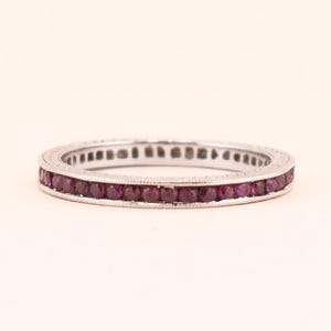 Peut inclure: Une bague en argent ornée d'une rangée continue de petites pierres précieuses rouge foncé de coupe carrée. La bague est posée sur un fond uni blanc cassé, mettant en valeur les pierres précieuses et le design de la bague. C'est un bijou.
