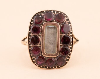 May include: A gold ring with a rectangular centre stone surrounded by a halo of small, dark red gemstones.