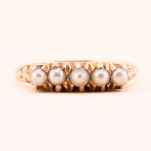 May include: A gold ring featuring five evenly spaced, round, white pearls. The ring has a textured band with a repeating pattern. The pearls are set in a row across the top of the ring.